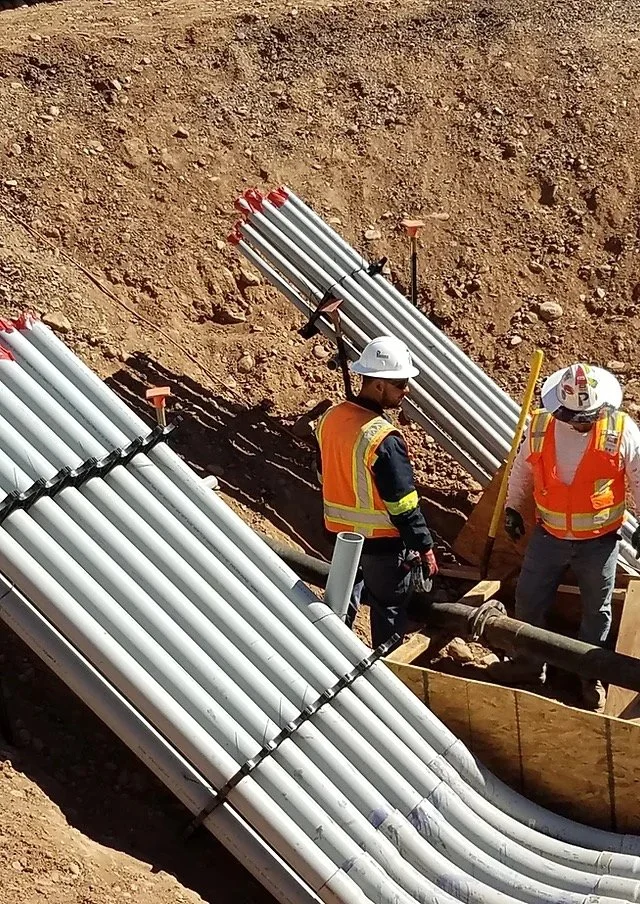 Workers inspect large white corrugated culvert pipes laid in excavated trench at construction site.